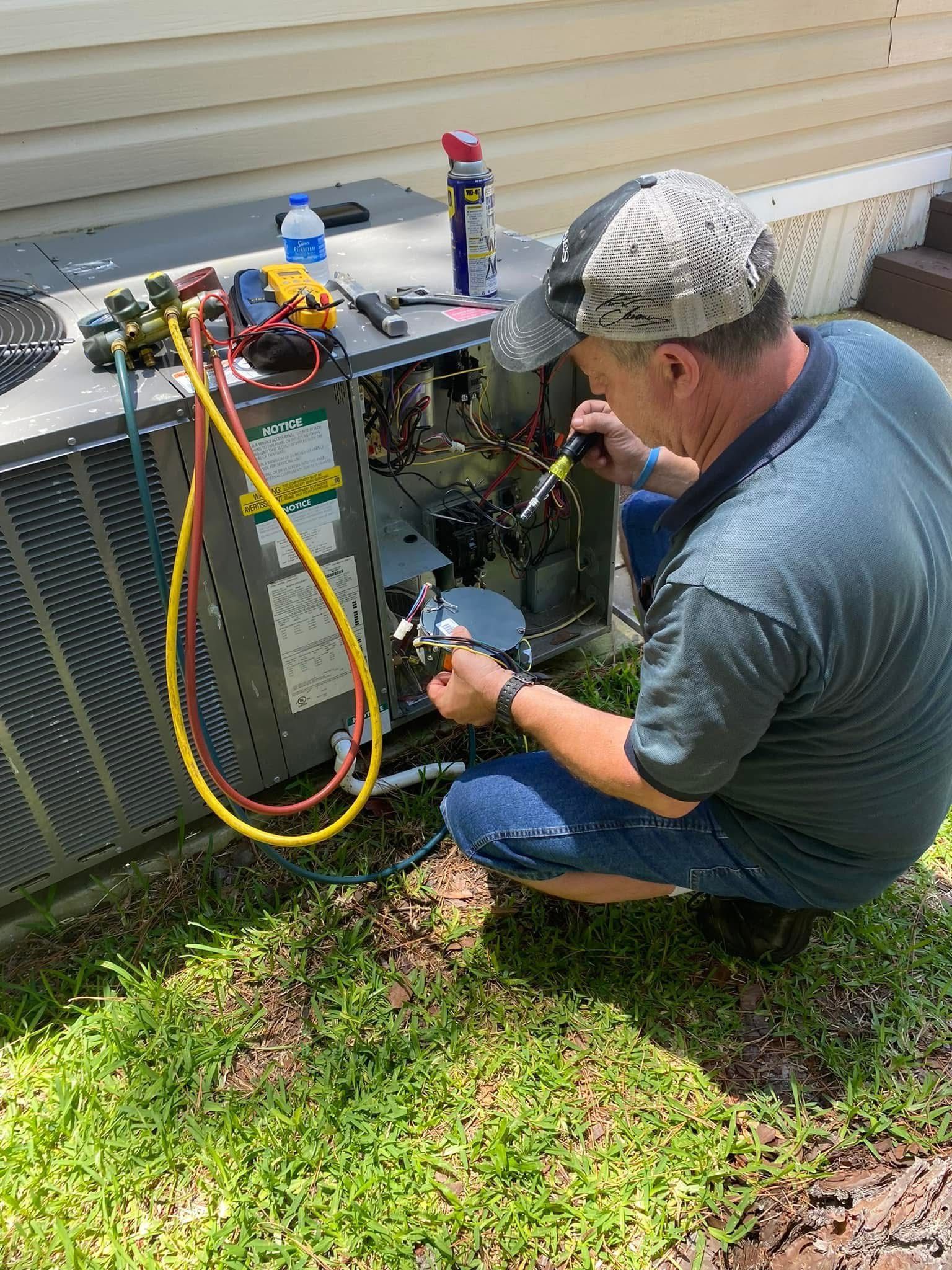 A man is kneeling down in the grass working on an air conditioner.