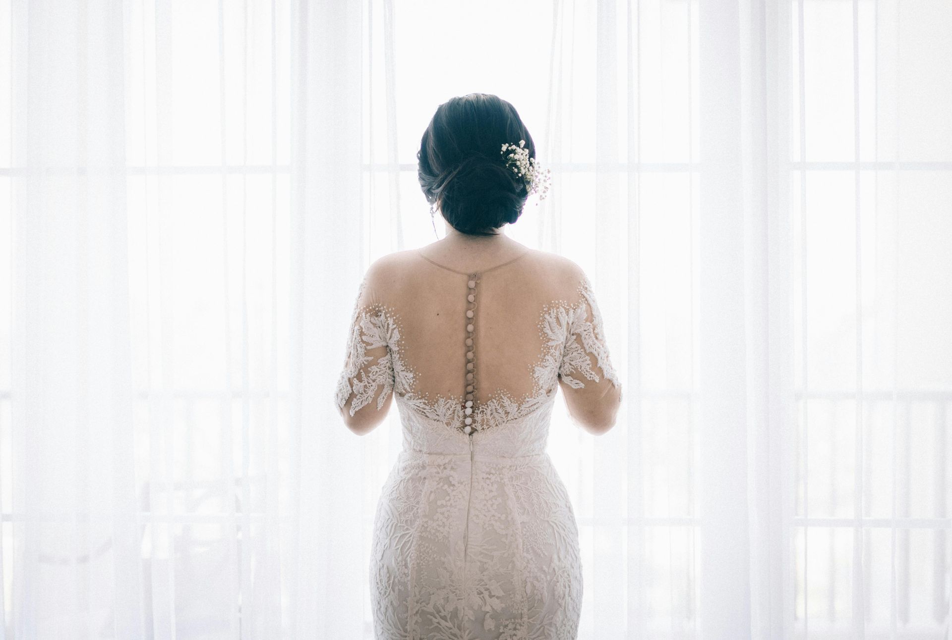 The back of a bride in a wedding dress is standing in front of a window.