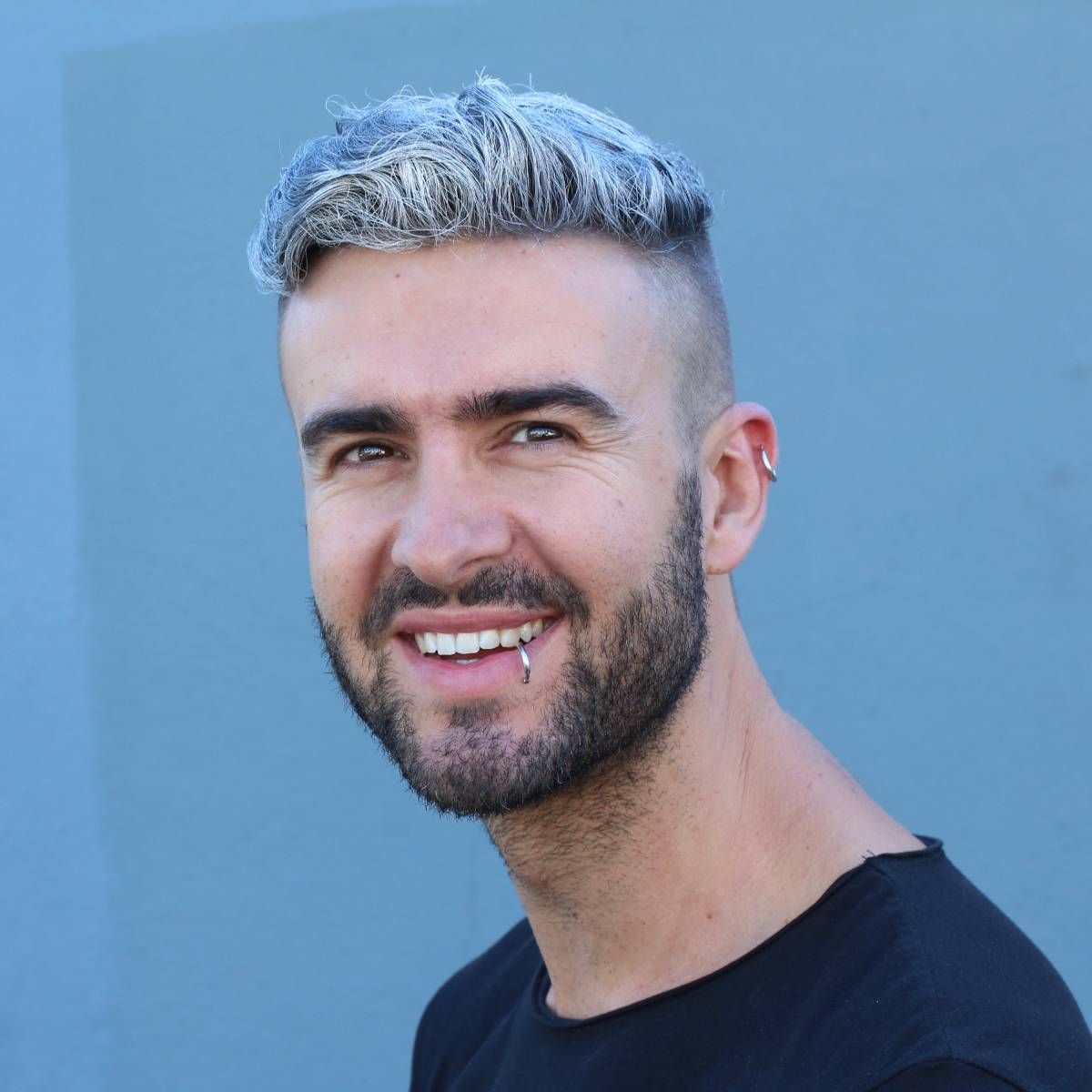 A man with a beard and piercings in his ears is smiling.