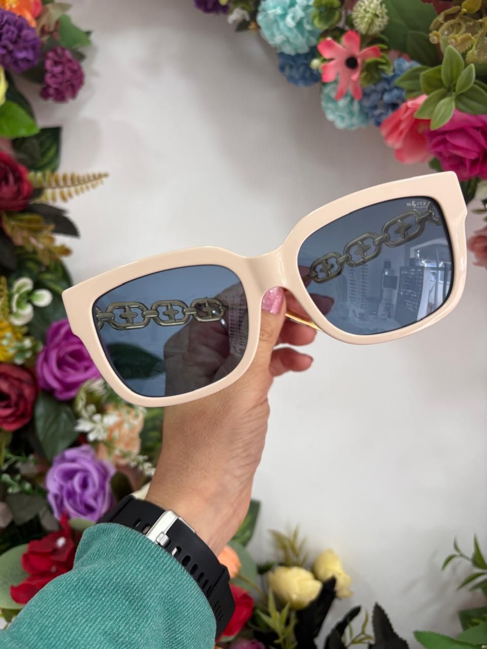 Mano sosteniendo unas gafas de sol de gran tamaño de color crema con lentes azules, cadena reflejada, contra un fondo floral.