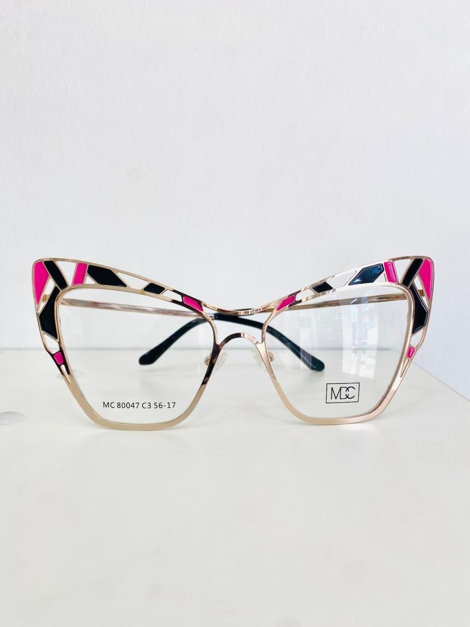 Gafas de ojo de gato con marco dorado y detalles estampados en rosa, negro y blanco sobre una superficie blanca.