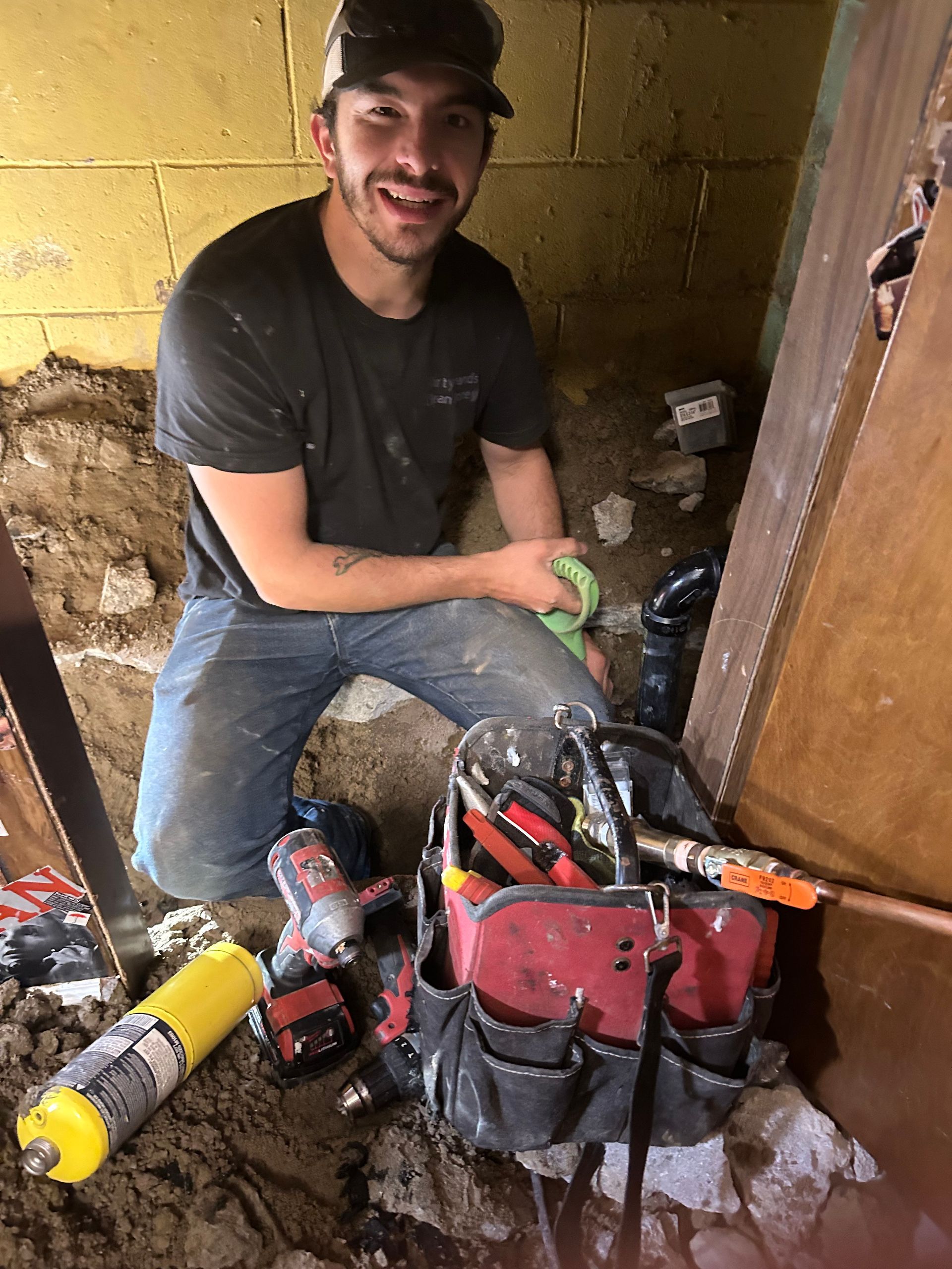 A man is kneeling down in the dirt next to a tool bag.