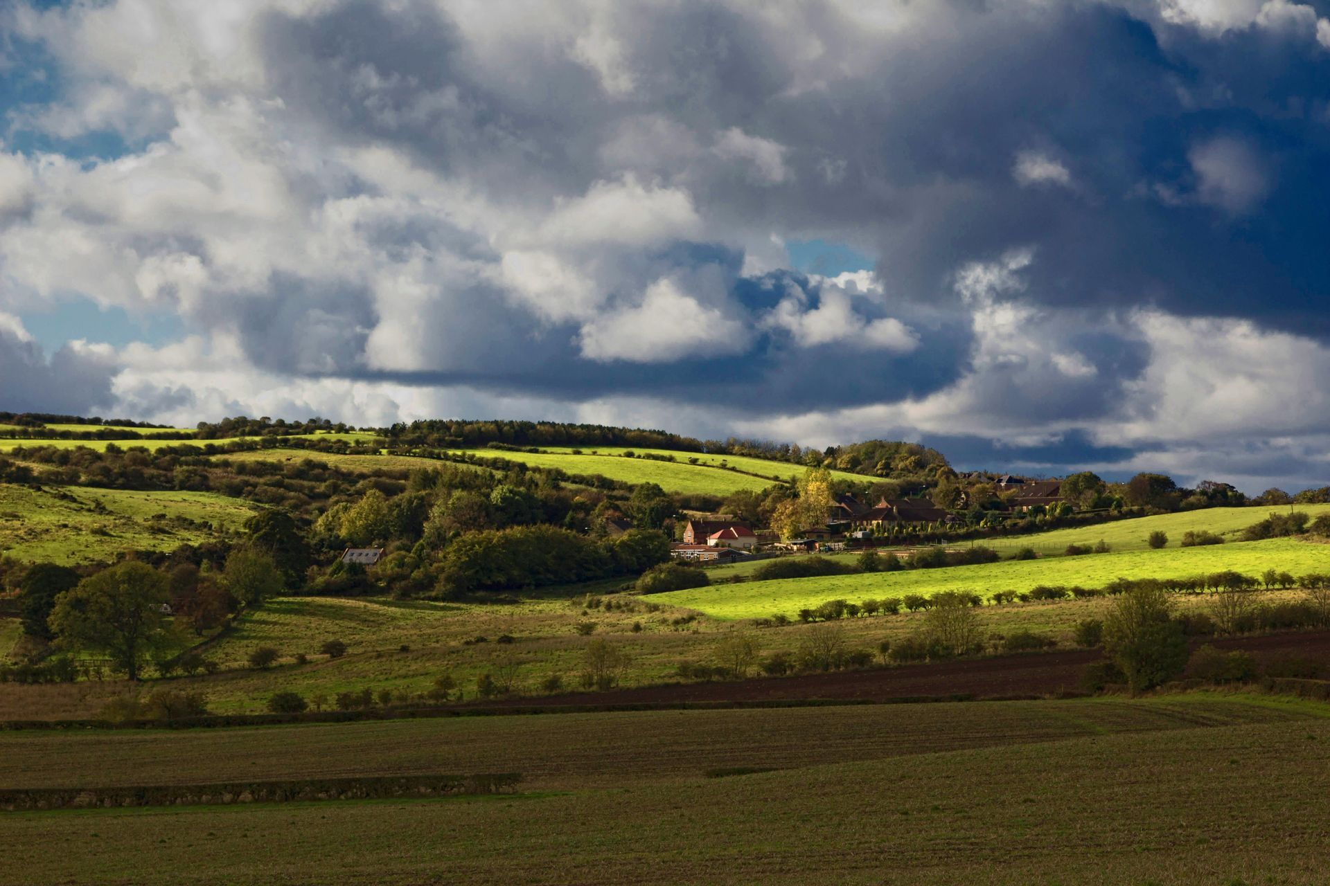 Rolling green hills under a dramatic cloudy sky, with a farmhouse nestled amongst trees.