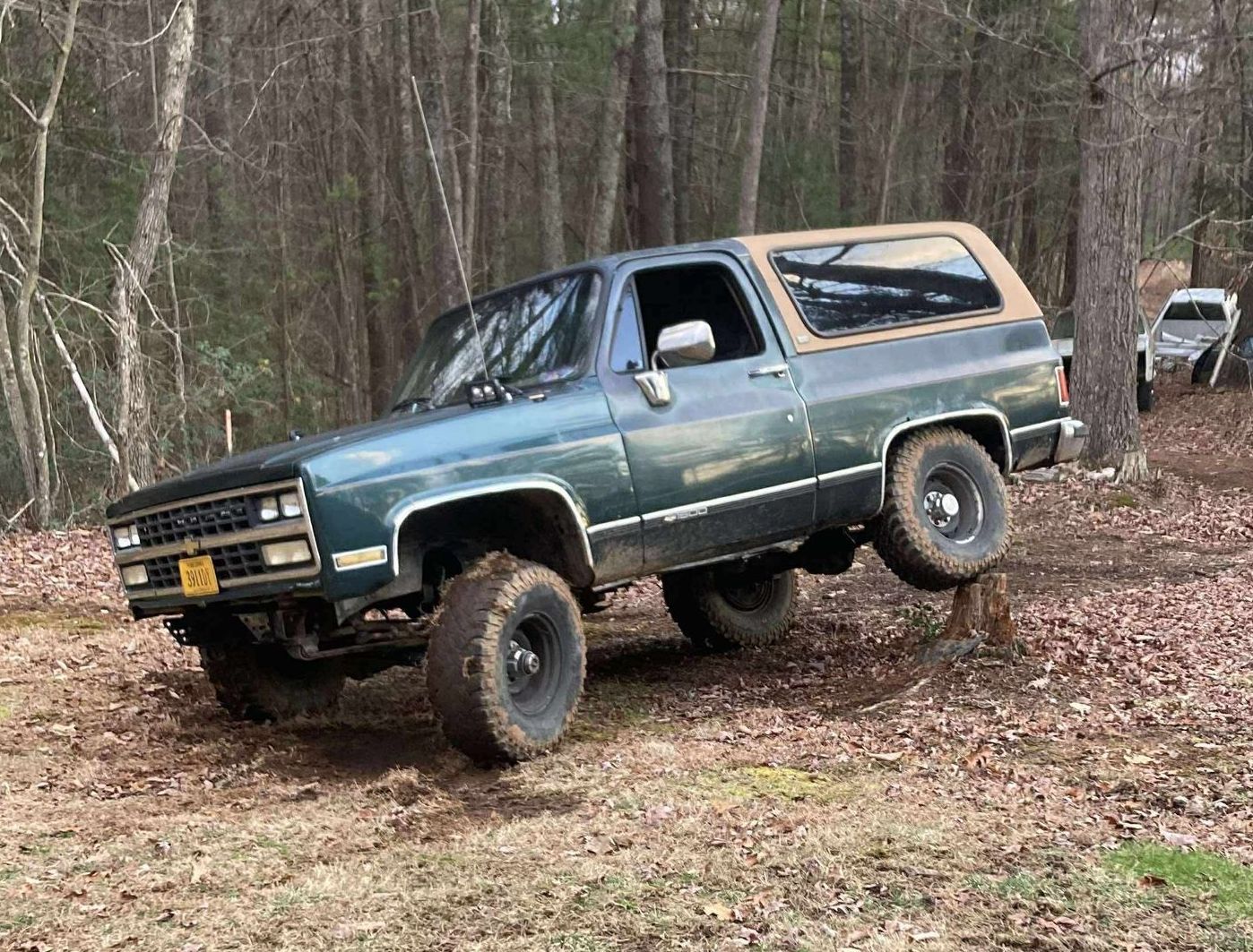 Green Blazer truck off-roading in a wooded area, front tire elevated on a stump.