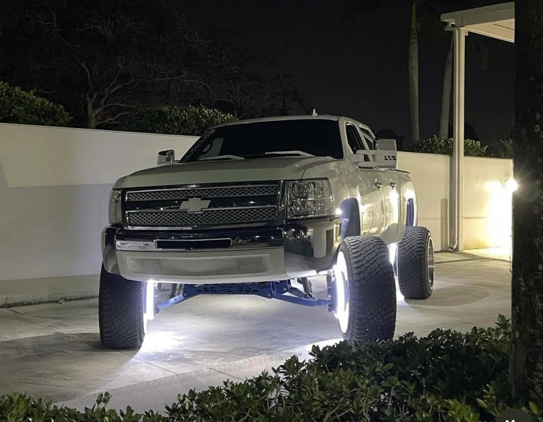 White lifted truck with blue underglow and rims, parked on a street at night.