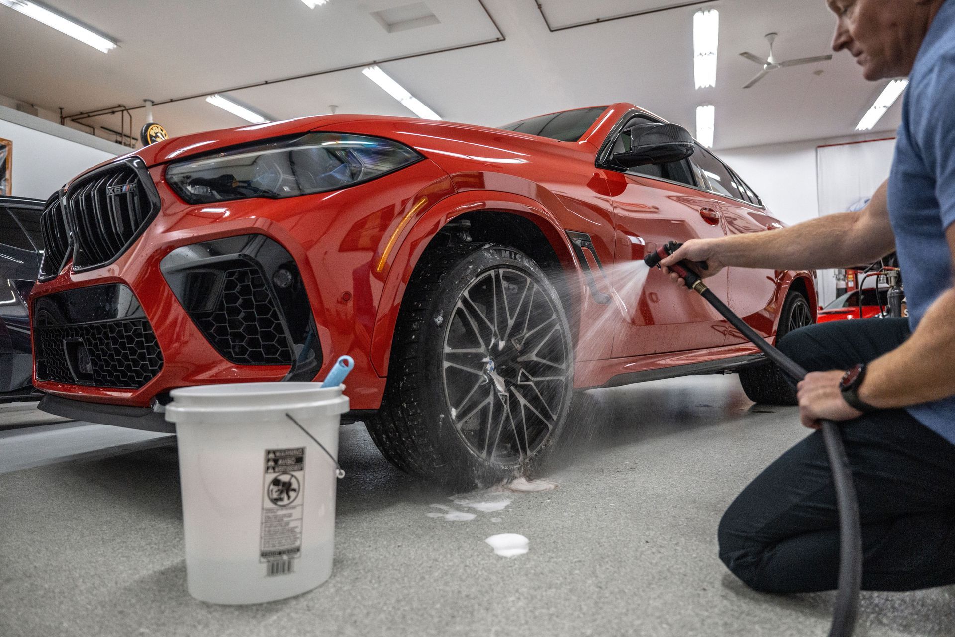 Person washing a red car in a garage, spraying water on the tire. A bucket sits nearby.