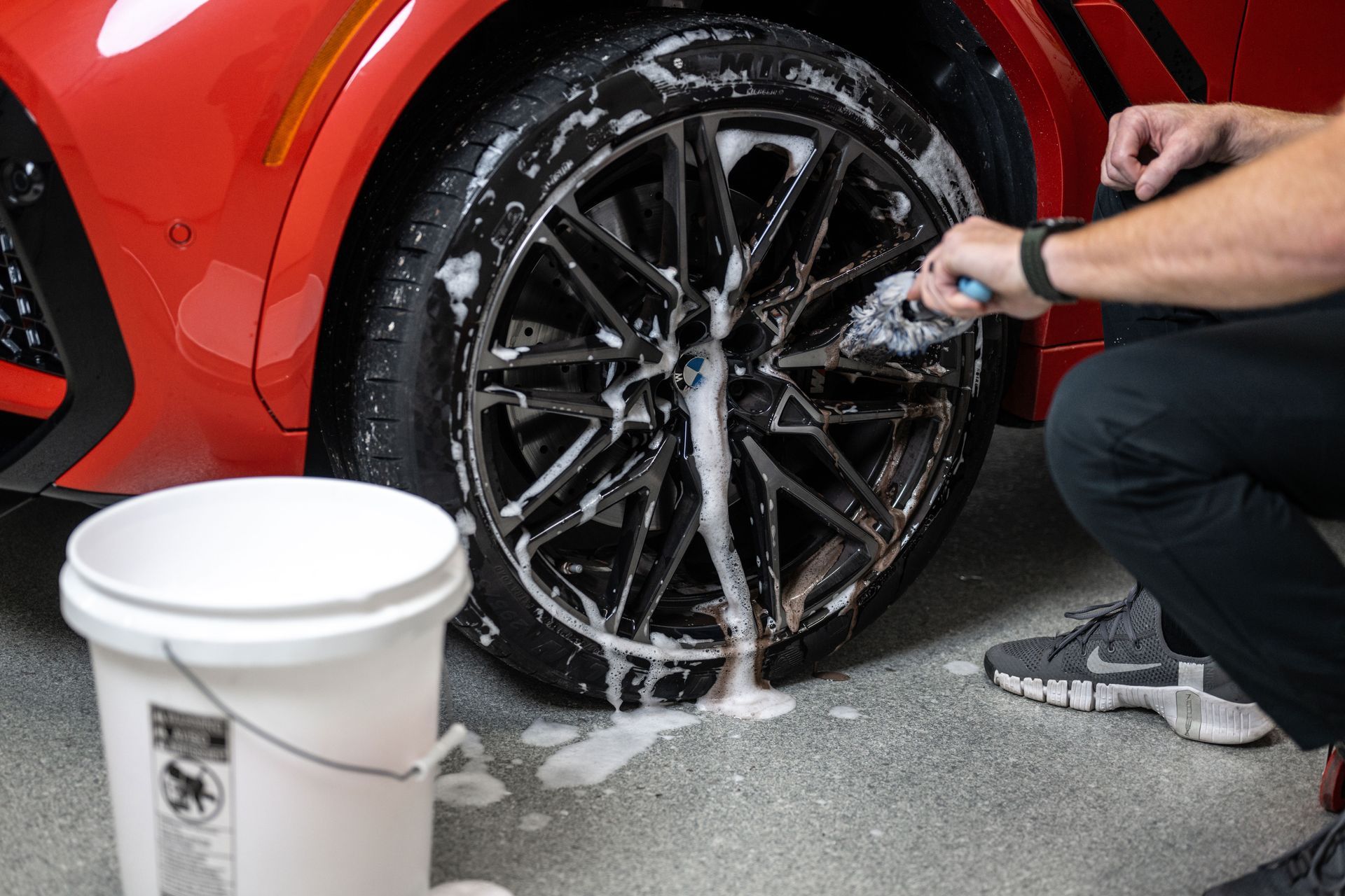 Person washing a black car wheel with soapy water, next to a red car and a bucket.