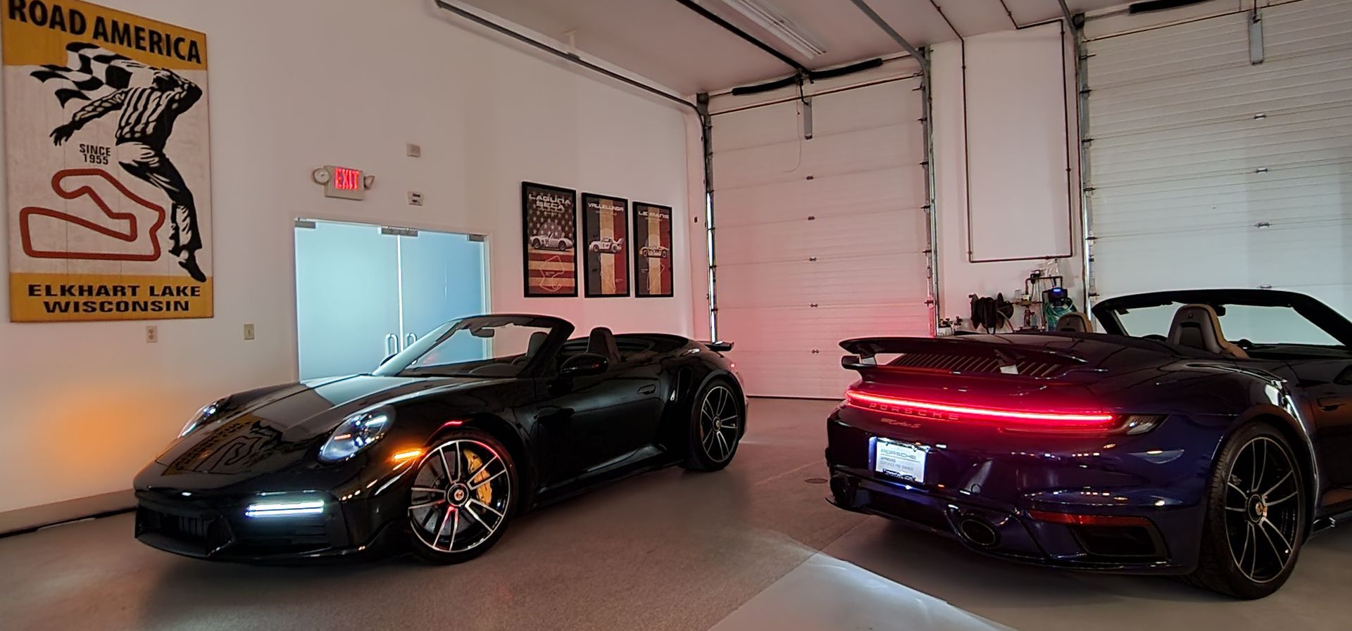 Two black convertibles parked in a garage, with a racing poster.