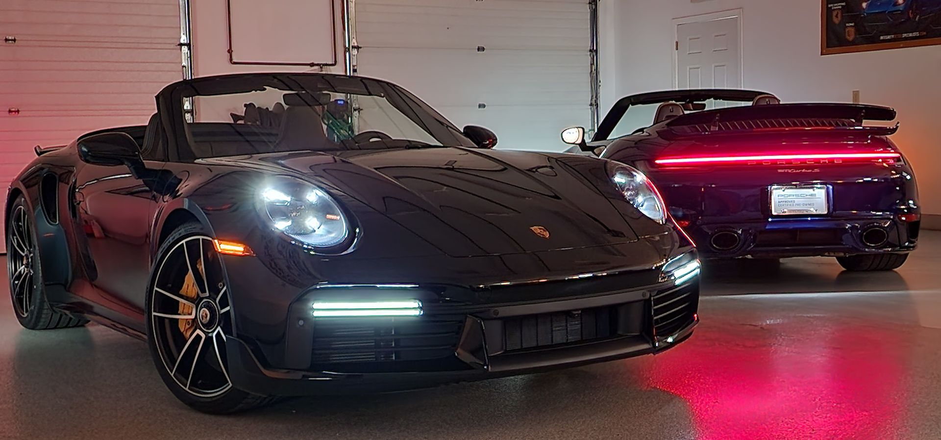 Two black Porsche 911s, one convertible, in a garage with red lighting.