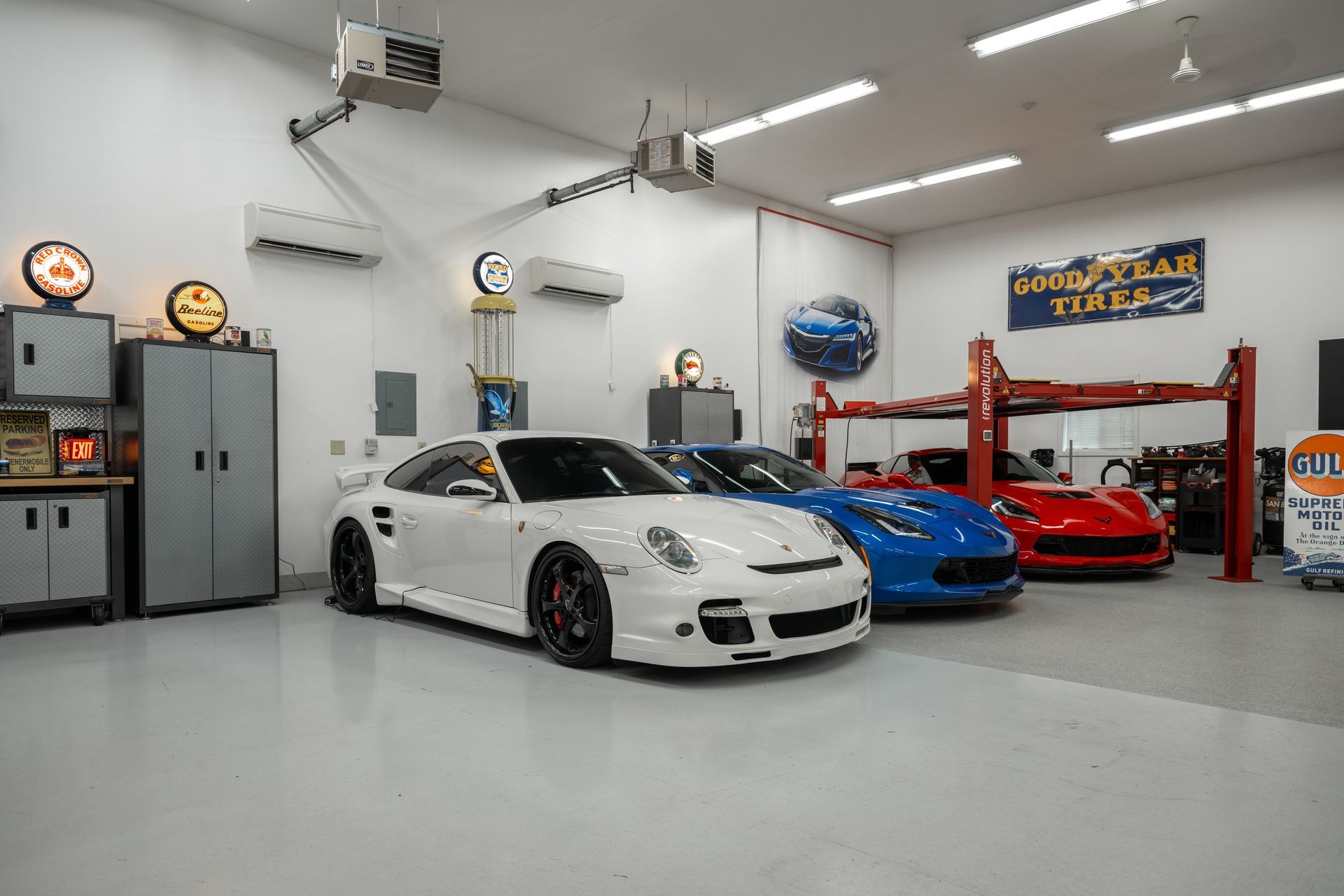 A spacious, well-lit garage with three sports cars: white, blue, and red. Includes a car lift and cabinets.