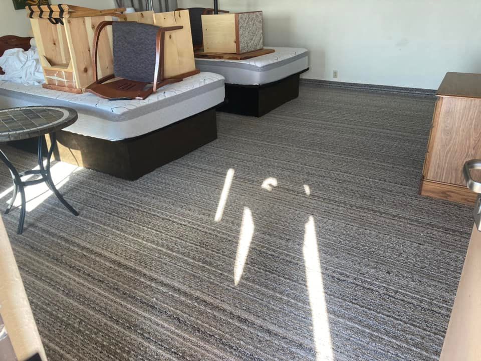 A cluttered room with mattresses, furniture, and carpeted flooring. Natural light streams through.