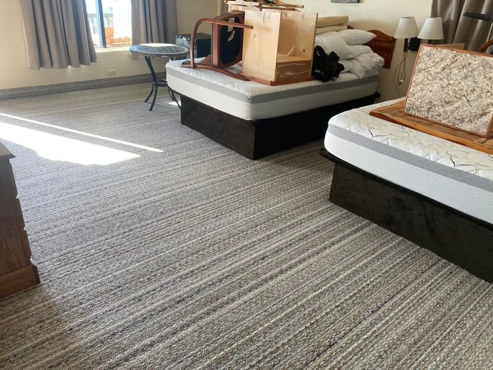 Two beds in a room with light brown carpet, boxes, and a small table near a window.