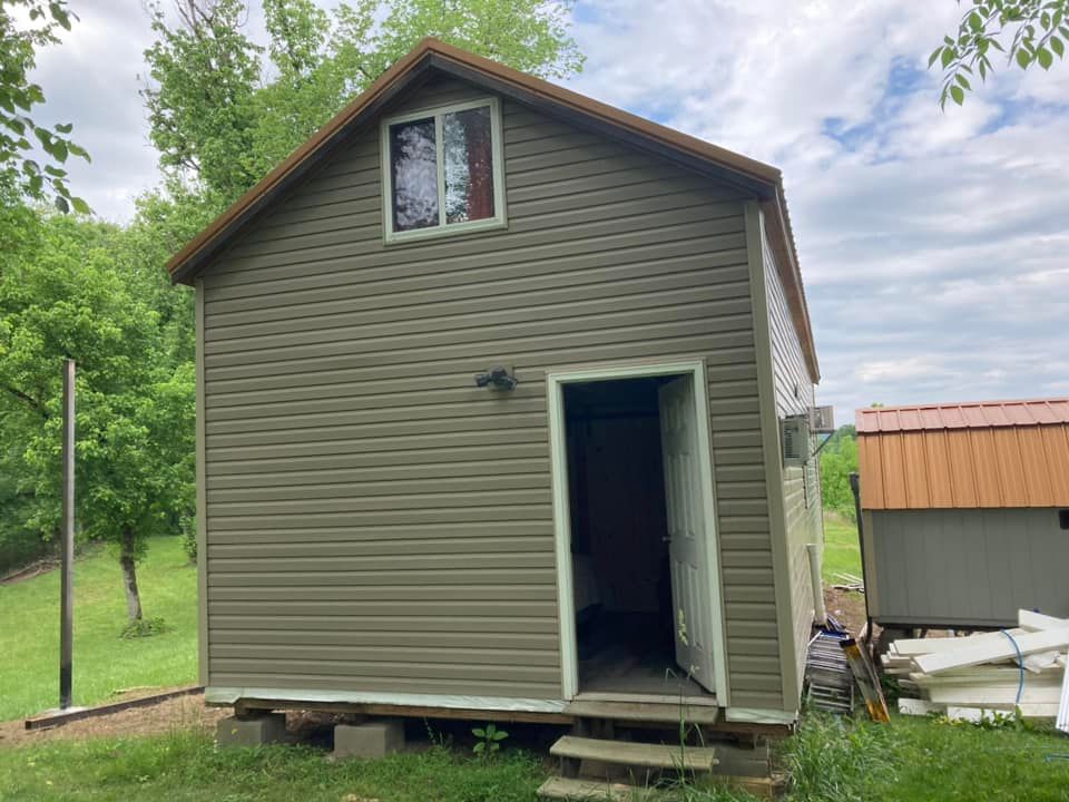 Green-sided two-story shed with open doorway, small window, and brown roof.
