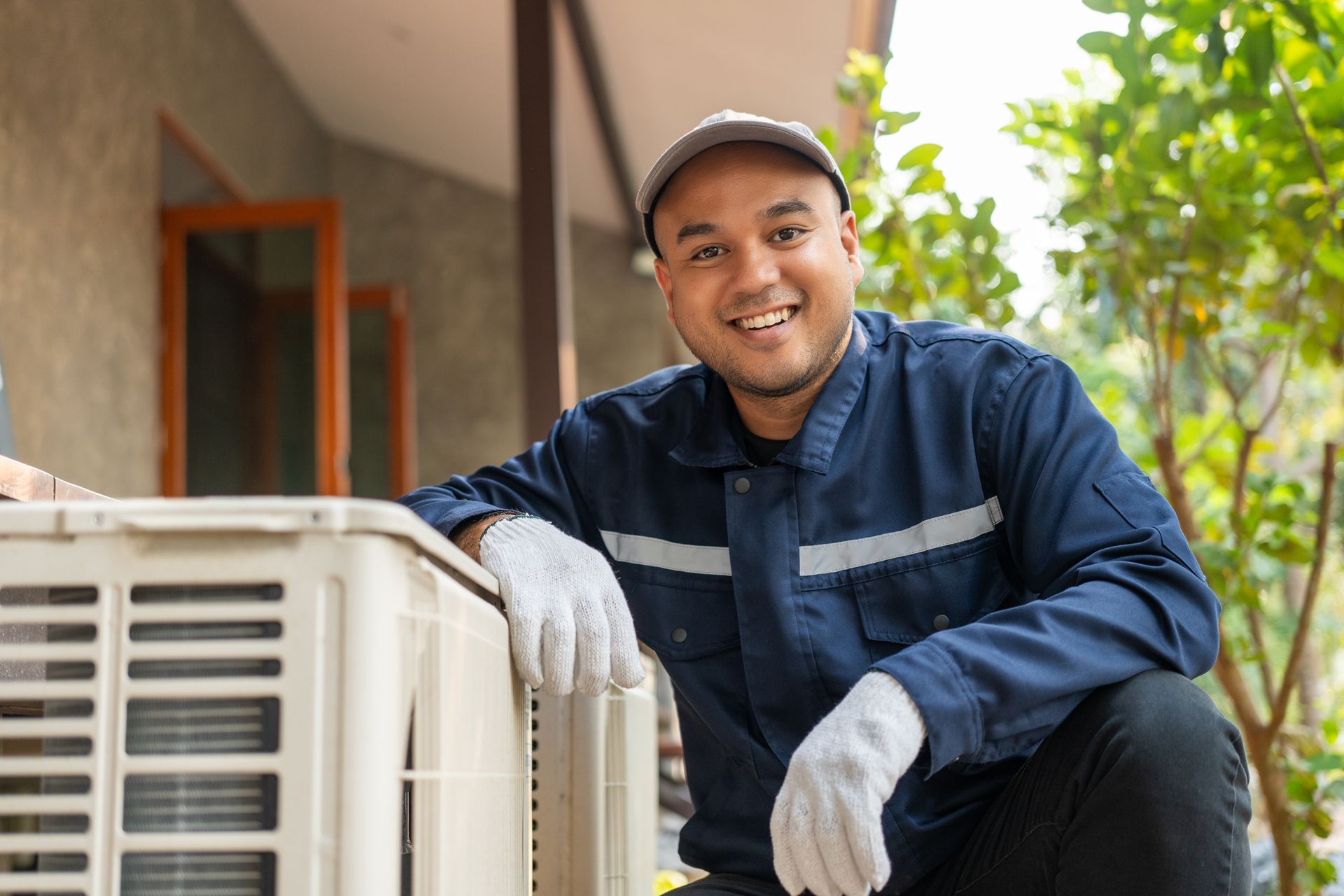 Man in blue work suit and gloves smiles near beige air conditioning unit.