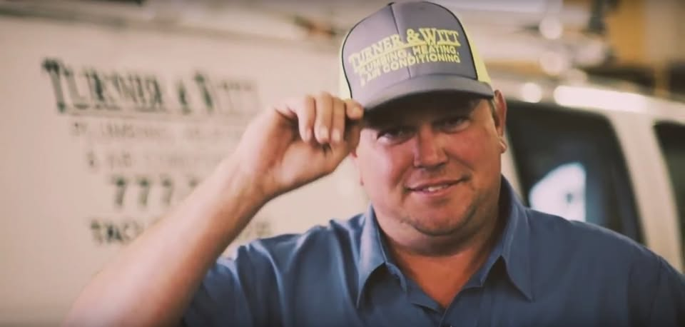 A man wearing a grey and yellow cap tips it. He smiles; text and a vehicle are in the background.
