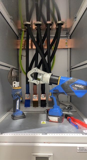 Two hydraulic crimping tools and a pair of pliers sit on the floor of an electrical cabinet with four heavy power cables.