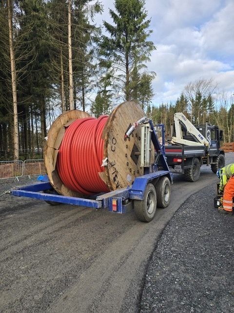 A large wooden reel of thick red cabling sits on a trailer attached to a truck on a gravel road by a forest.