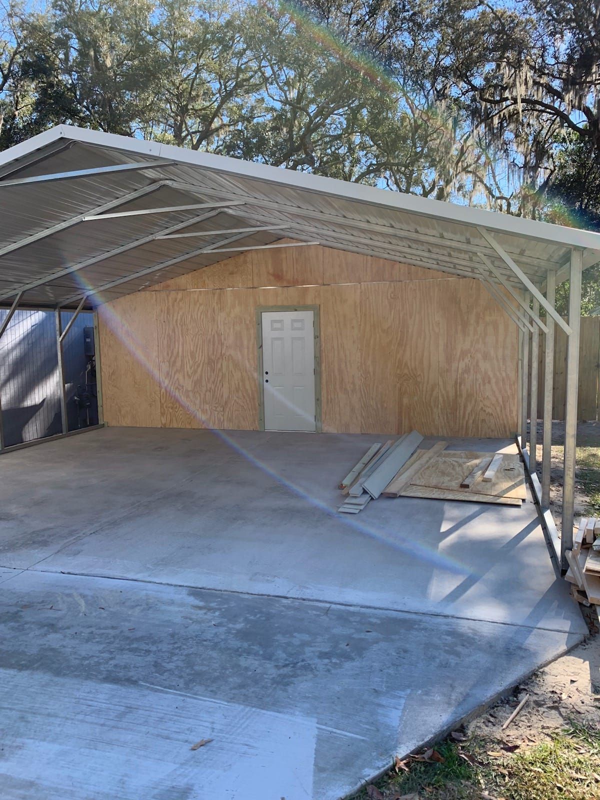 Carport with concrete floor and plywood wall, white door. Metal frame, outdoors.
