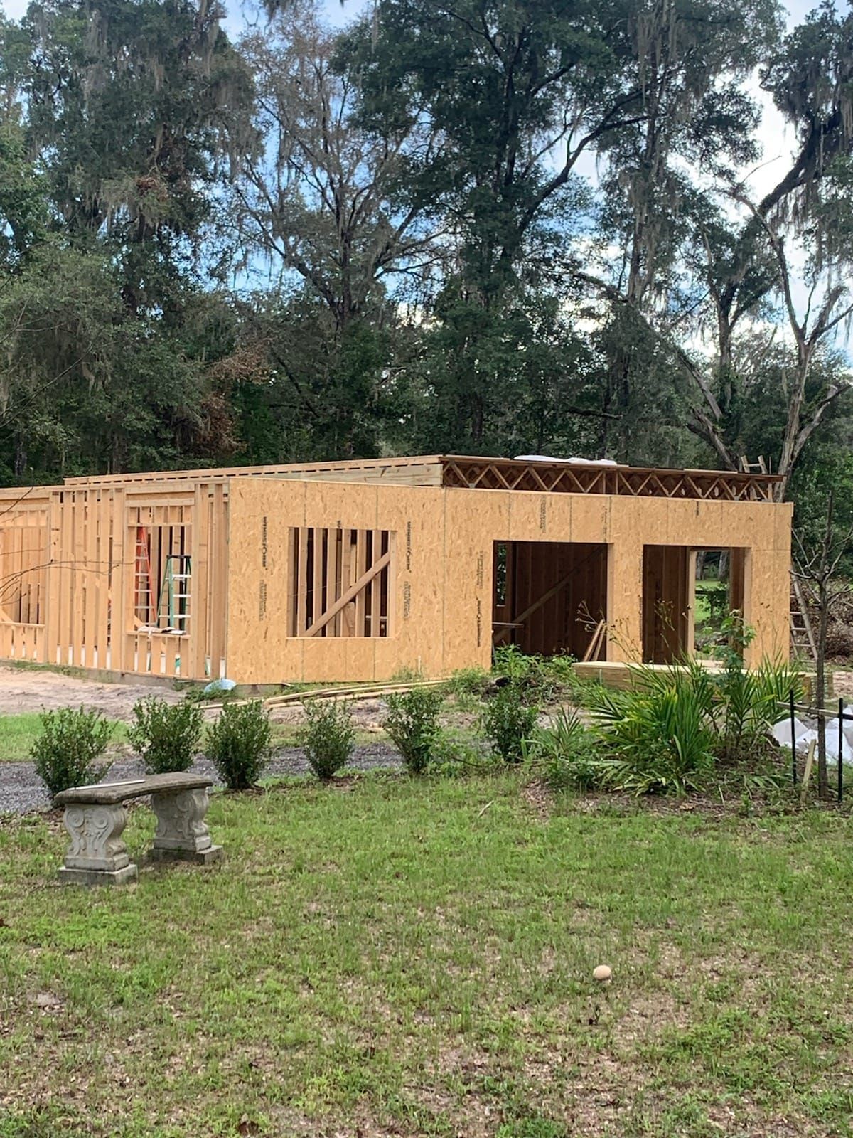 A wood-framed building under construction in a grassy yard, with a garden bench in the foreground and trees in the background.