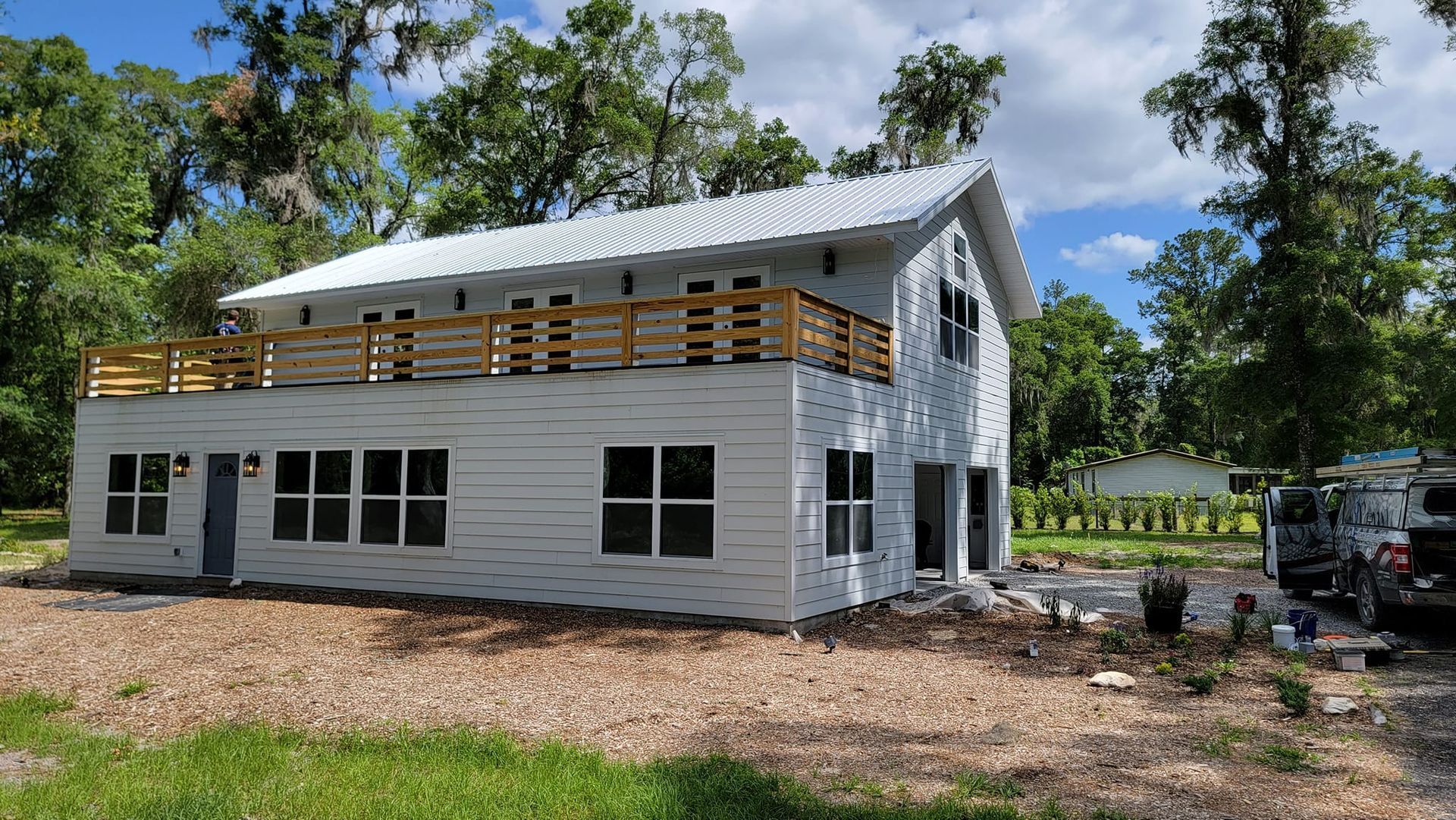 White two-story house with a balcony, metal roof, and windows surrounded by trees and landscaping.