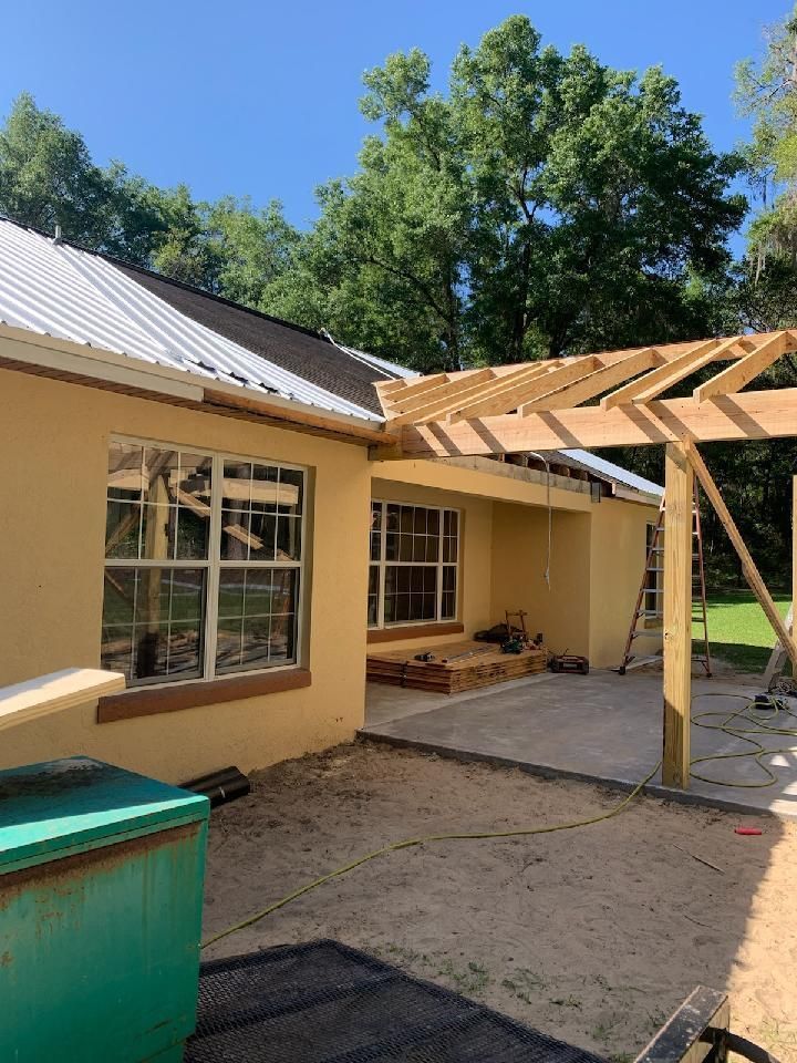 Yellow house with a metal roof, under construction. A wooden pergola is being built over a concrete patio.
