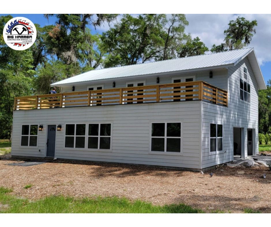 White two-story house with metal roof, wood deck, and large windows, set among trees and grass.