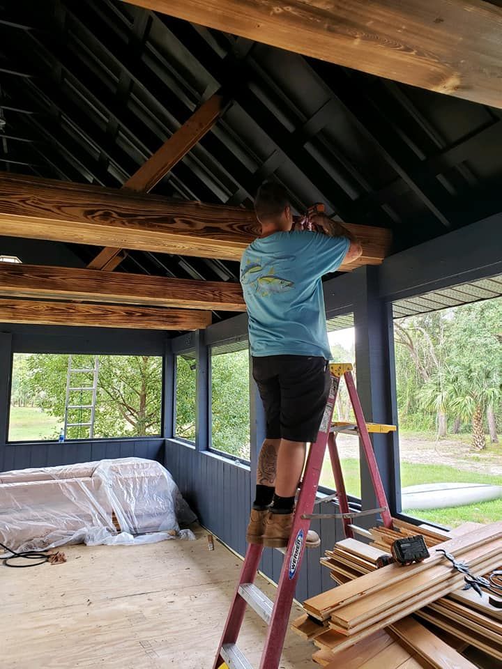 Man on ladder installing wood beams in a room with blue walls and large windows.