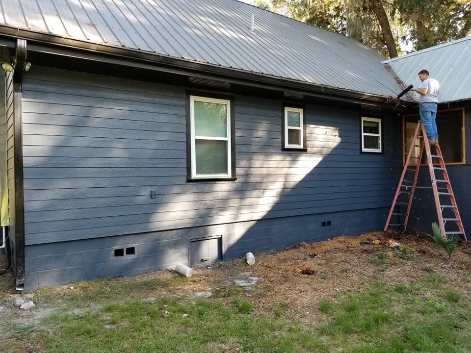Man on a ladder cleaning gutters of a blue house with white-framed windows.