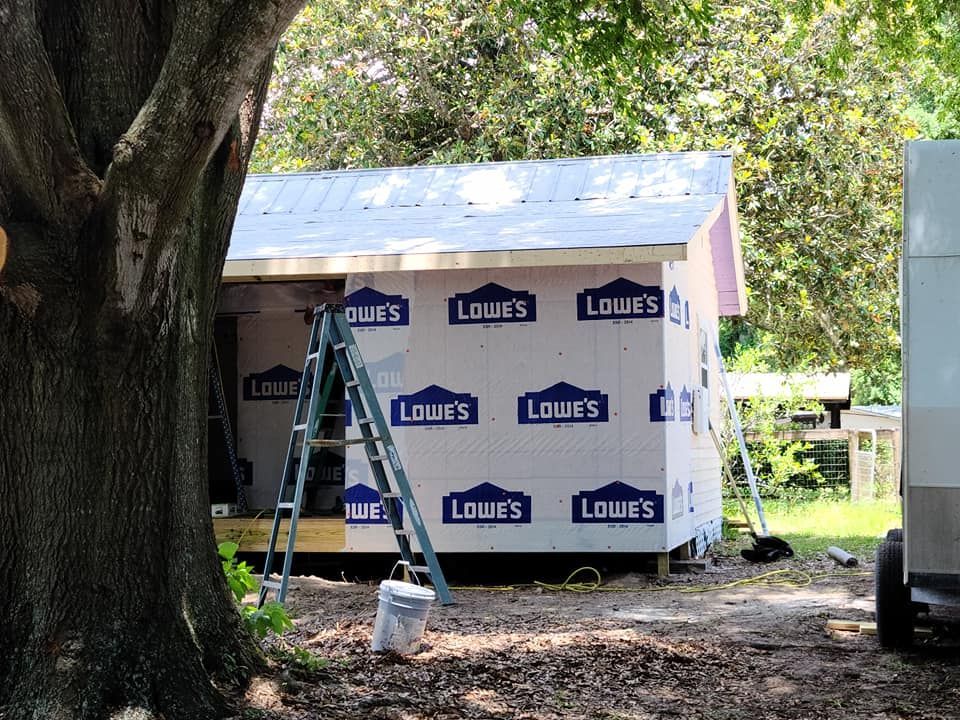 Construction of a small shed. Lowe's insulation covers walls, a ladder leans against it. Tree in foreground.