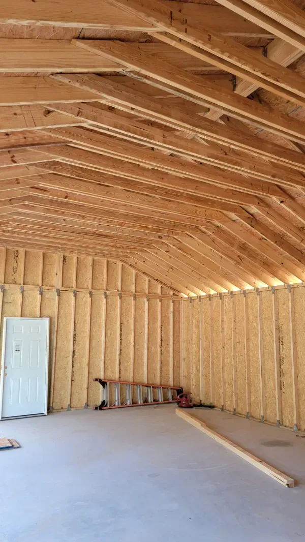 Interior of a building under construction, showing exposed wooden rafters and studs.  A door, ladder, and lumber are visible.