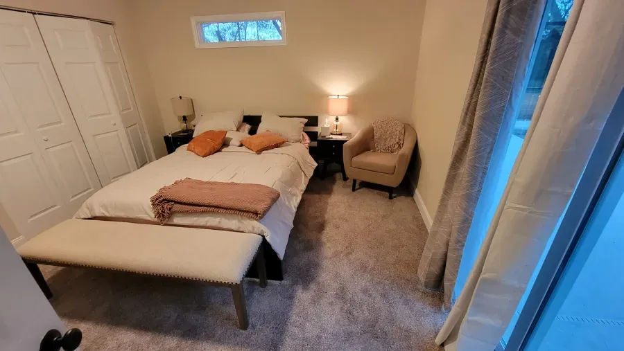 Bedroom with bed, bench, nightstands, armchair, and closet. Beige and orange accents.