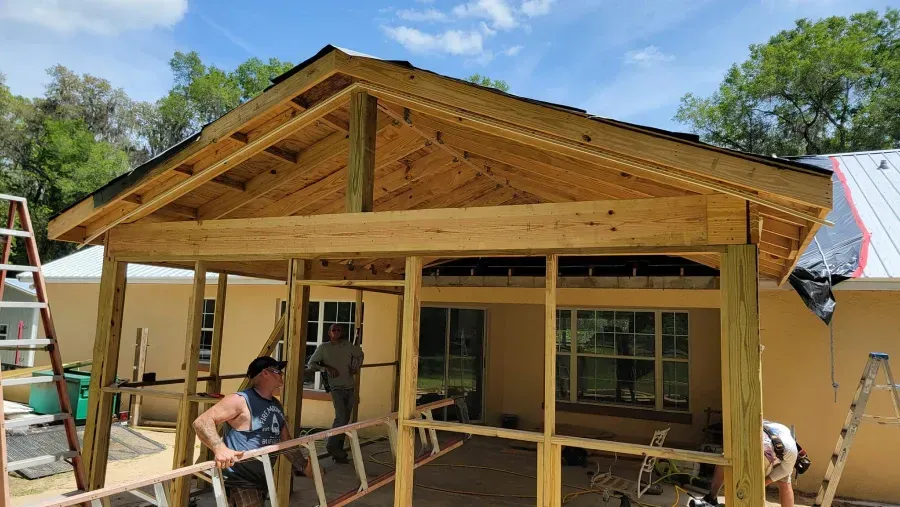 Construction of a wooden covered patio attached to a beige house; workers carrying ladders, sunny outdoors.