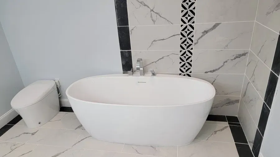A white oval bathtub and toilet in a bathroom with marble tile, black accents, and light blue walls.