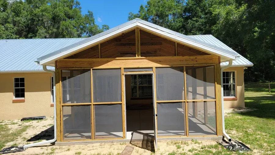 Screened porch with wooden frame and metal roof. Open screen door, backyard setting.