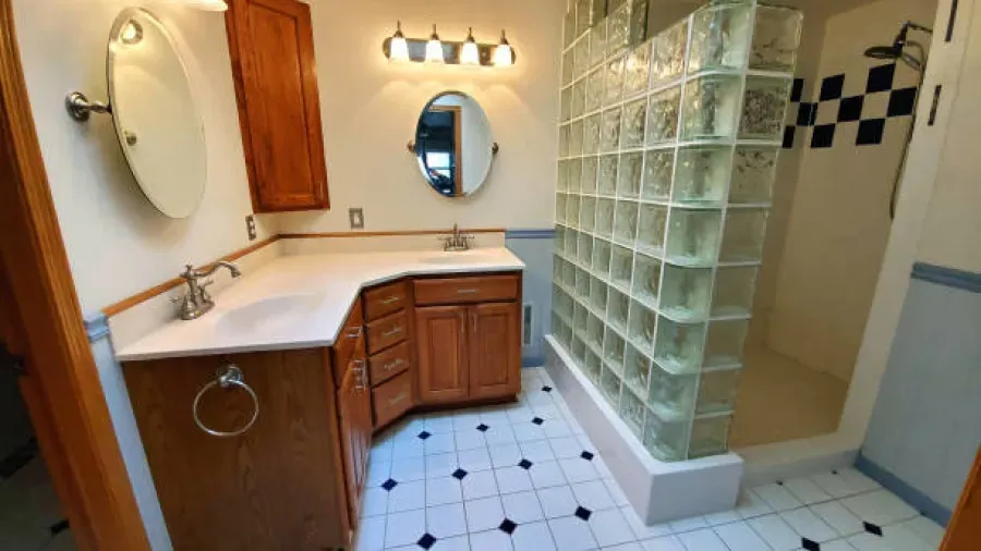 Bathroom with light wood cabinets, white countertops, and a glass block shower. Black and white tiled floor.