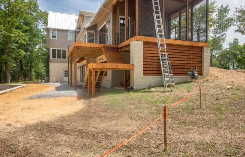 Construction site: house with unfinished deck, screened porch, and ladder, dirt foreground.