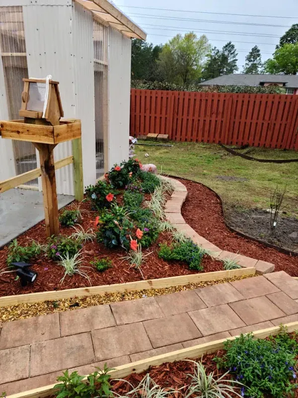 A garden with a path, flower beds, and a bird feeder next to a white shed, with a red fence in the background.