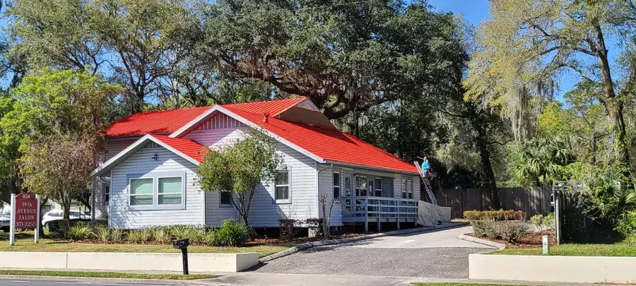 A light blue house with a bright red roof sits amongst trees, with a small sign in front of the house.