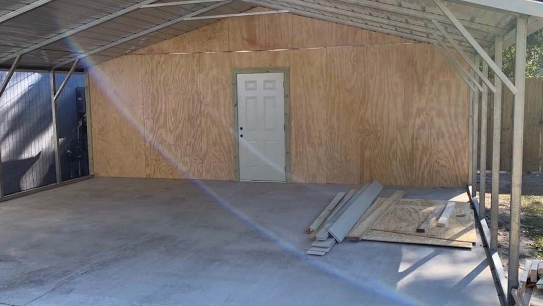 Concrete floored carport with plywood walls and a white door.
