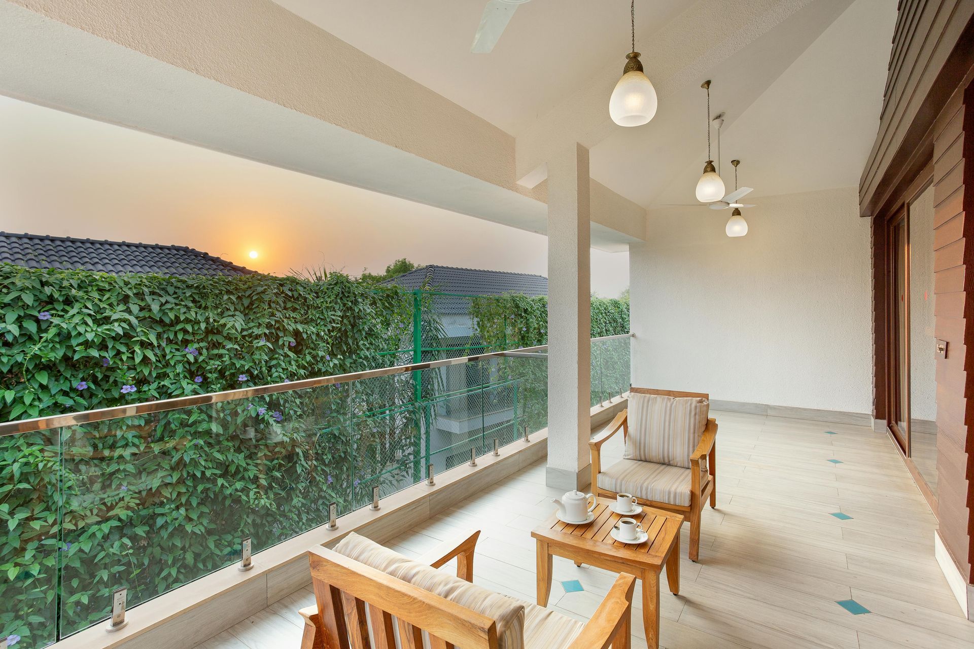 Balcony with wooden chairs, a small table with tea cups, and a green hedge with sunset in the background.