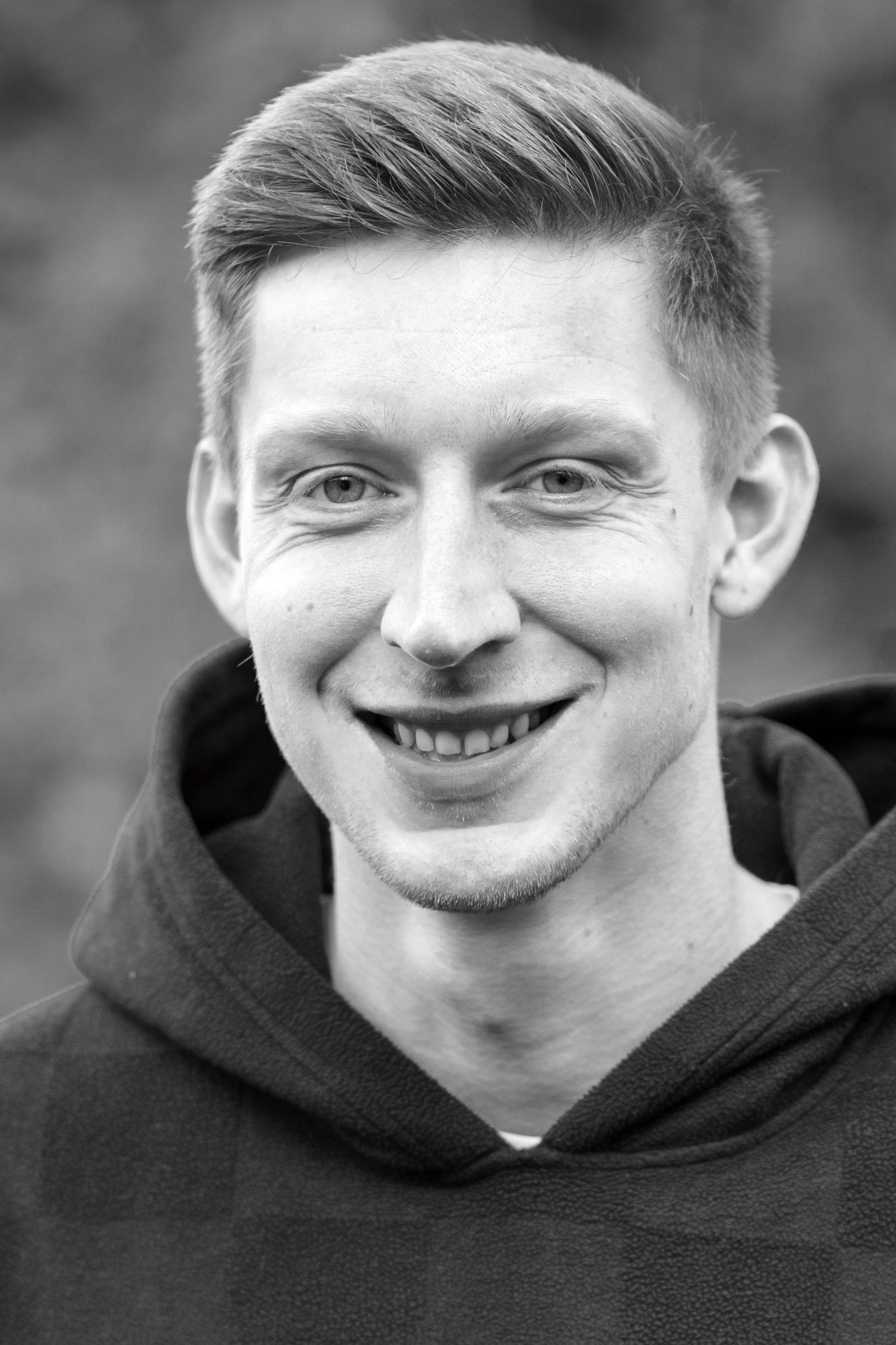A black and white photo of a smiling young man wearing a hoodie.
