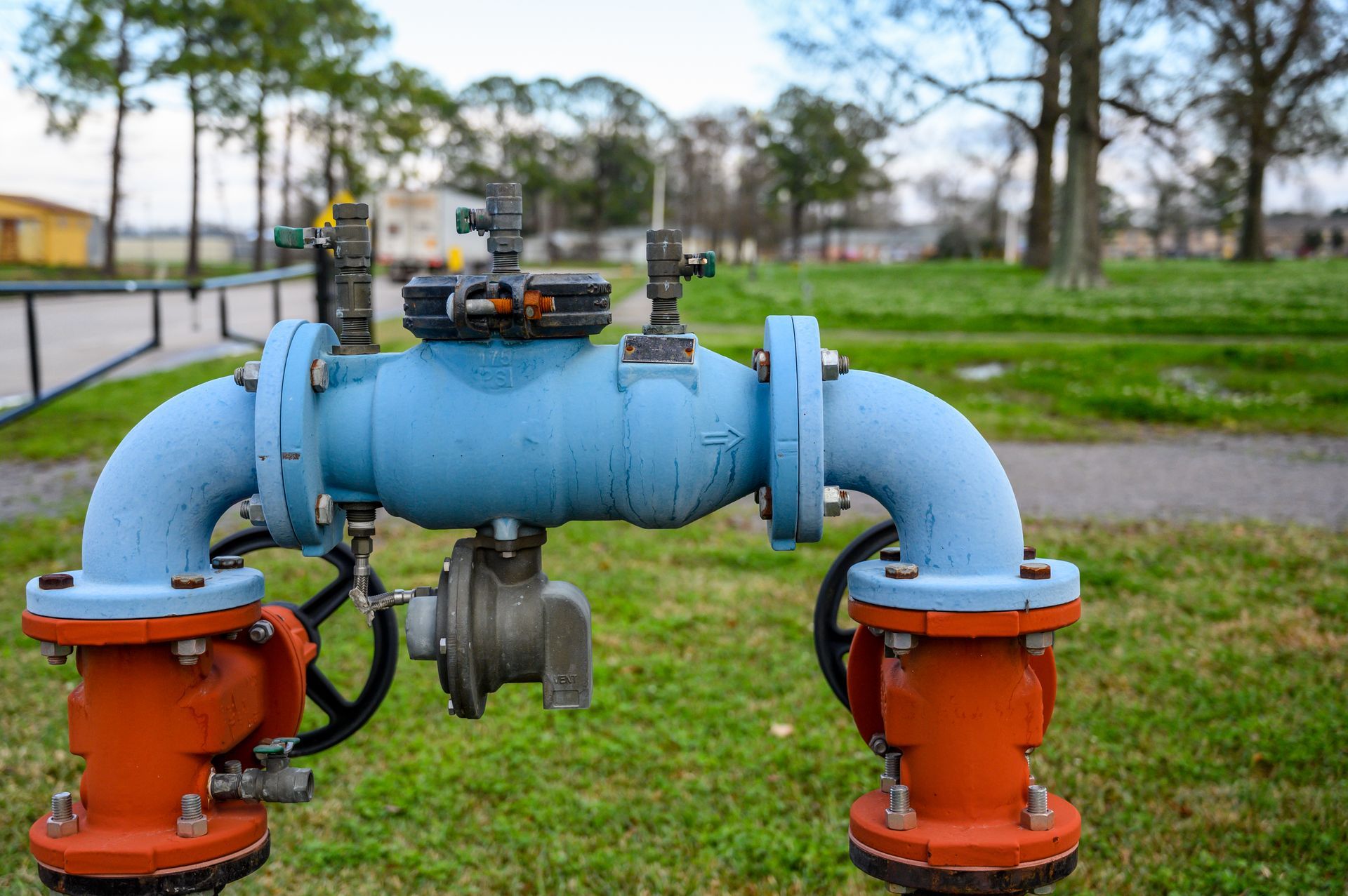 Blue and orange water valve on green grass with trees and buildings in the background.
