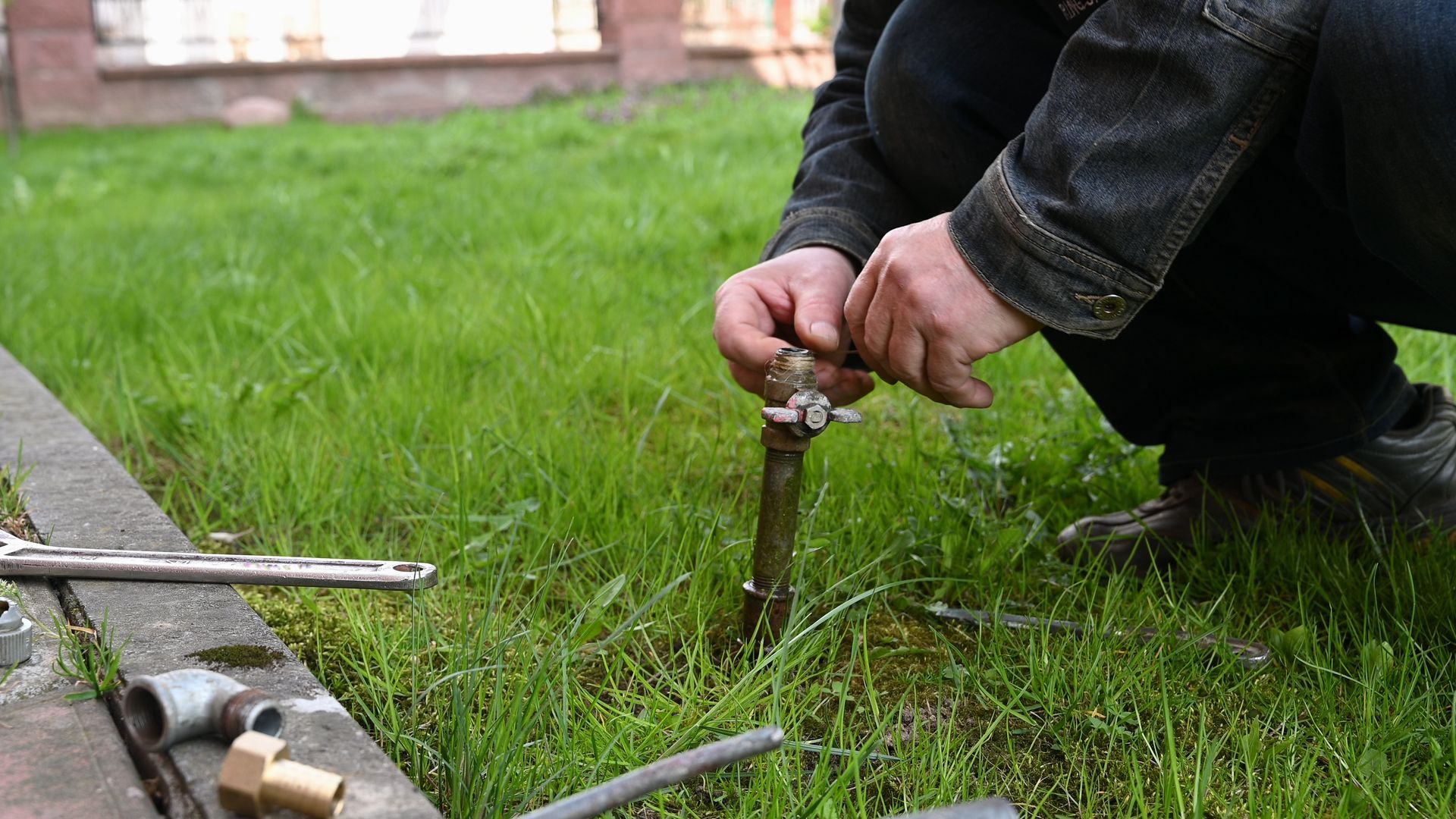 A man is kneeling down in the grass fixing a faucet.
