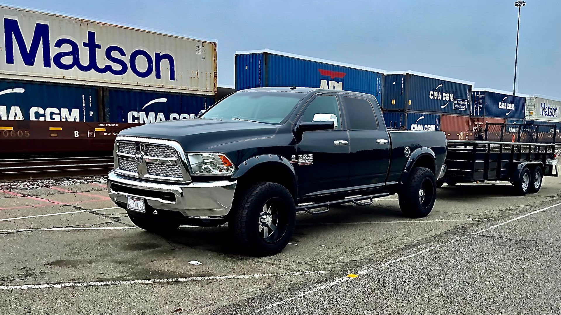 Black pickup truck with trailer parked in a lot in front of freight railcars and a Matson container.
