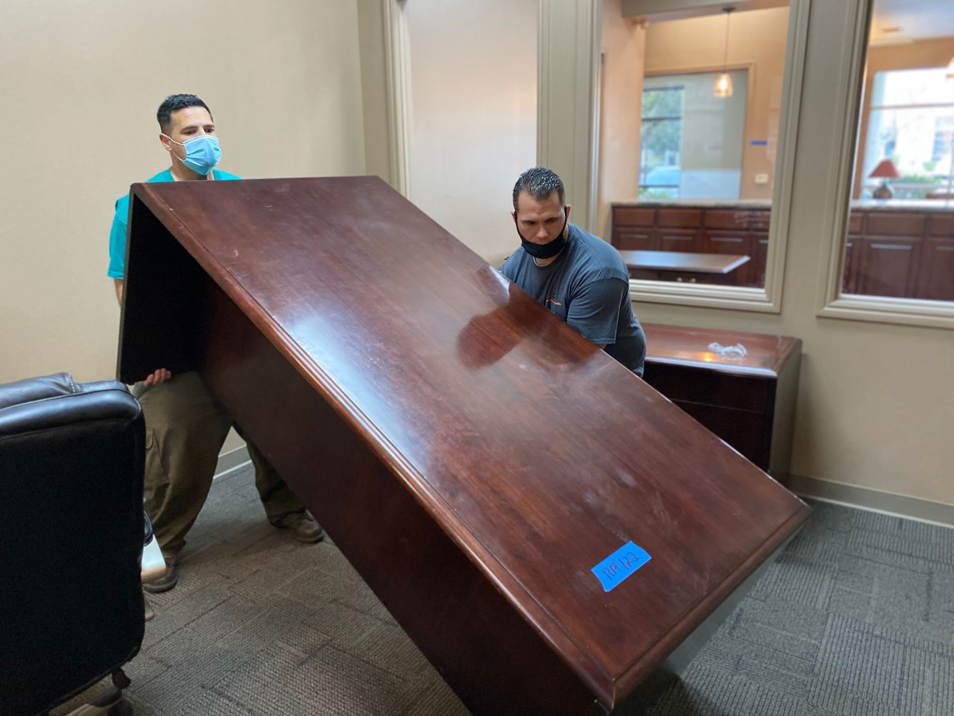 Two people carry a large dark wooden panel through an office hallway.