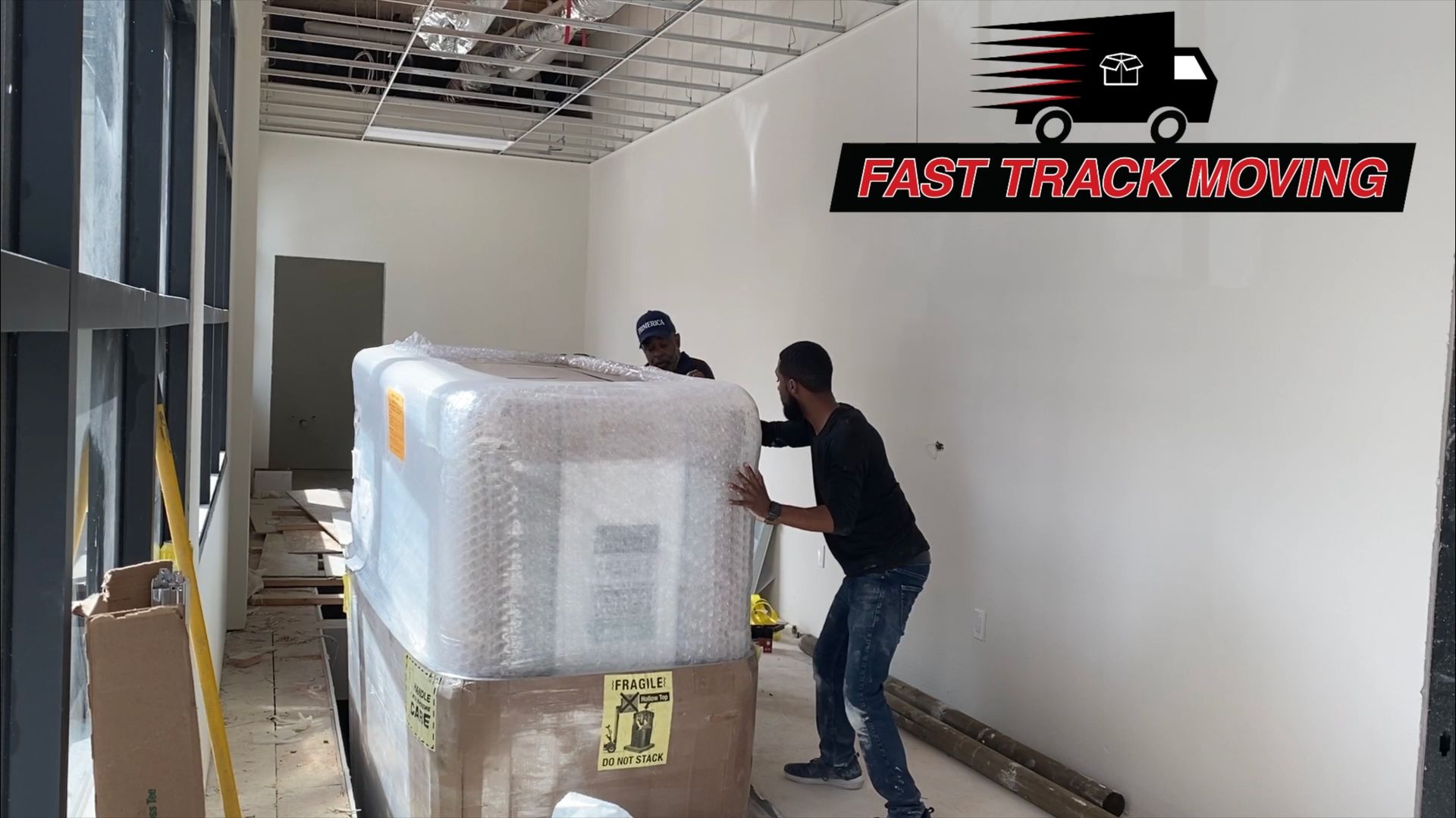 Two movers pushing a wrapped pallet through a narrow hallway inside a building.