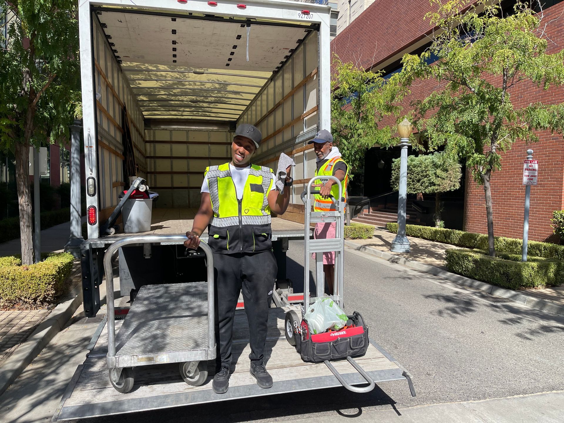 Two utility workers in high-visibility vests beside an open box truck on a driveway, with equipment and a cart.