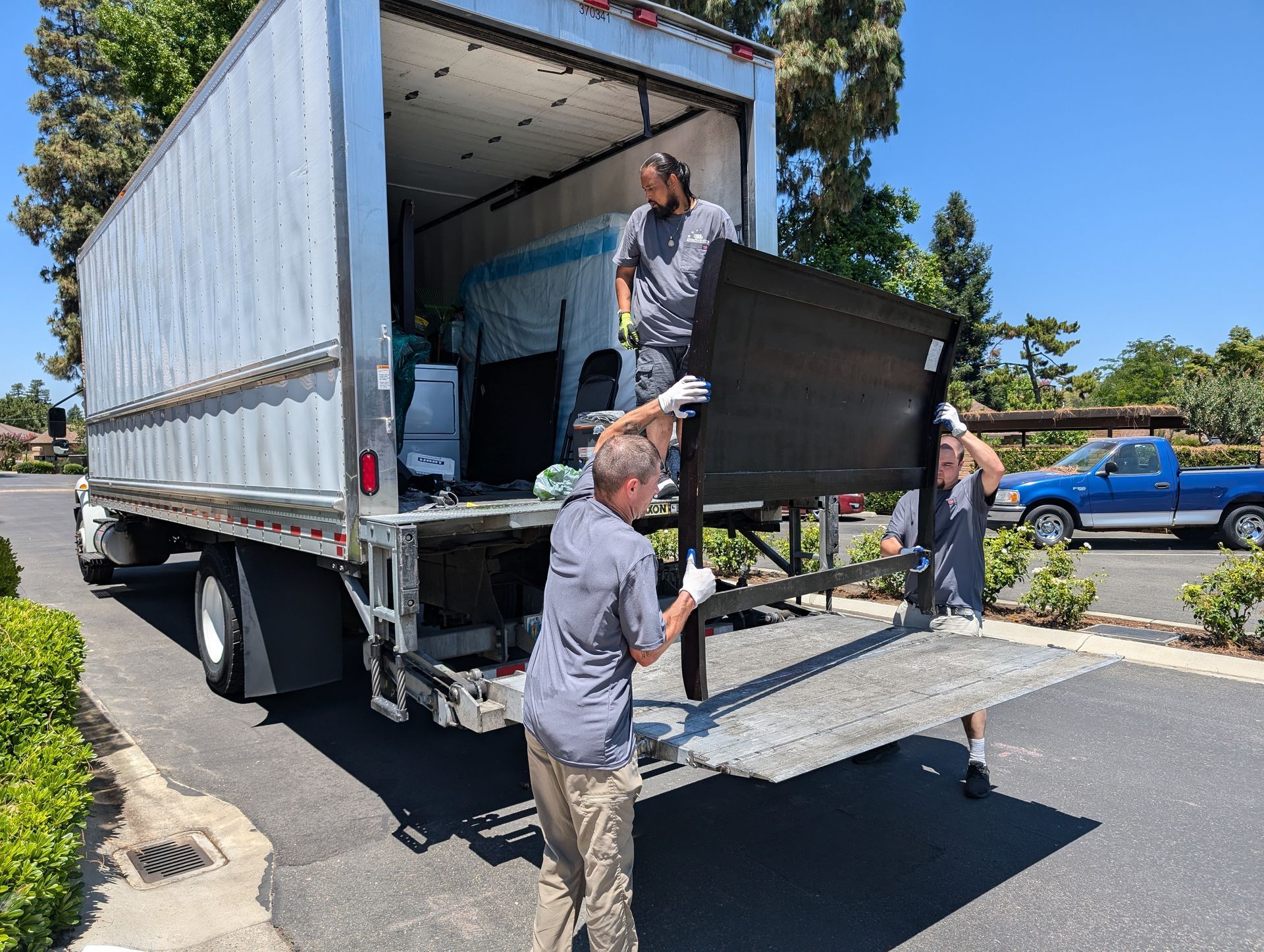 Workers unloading furniture from a moving truck on a sunny street, lowering a ramp at the back.