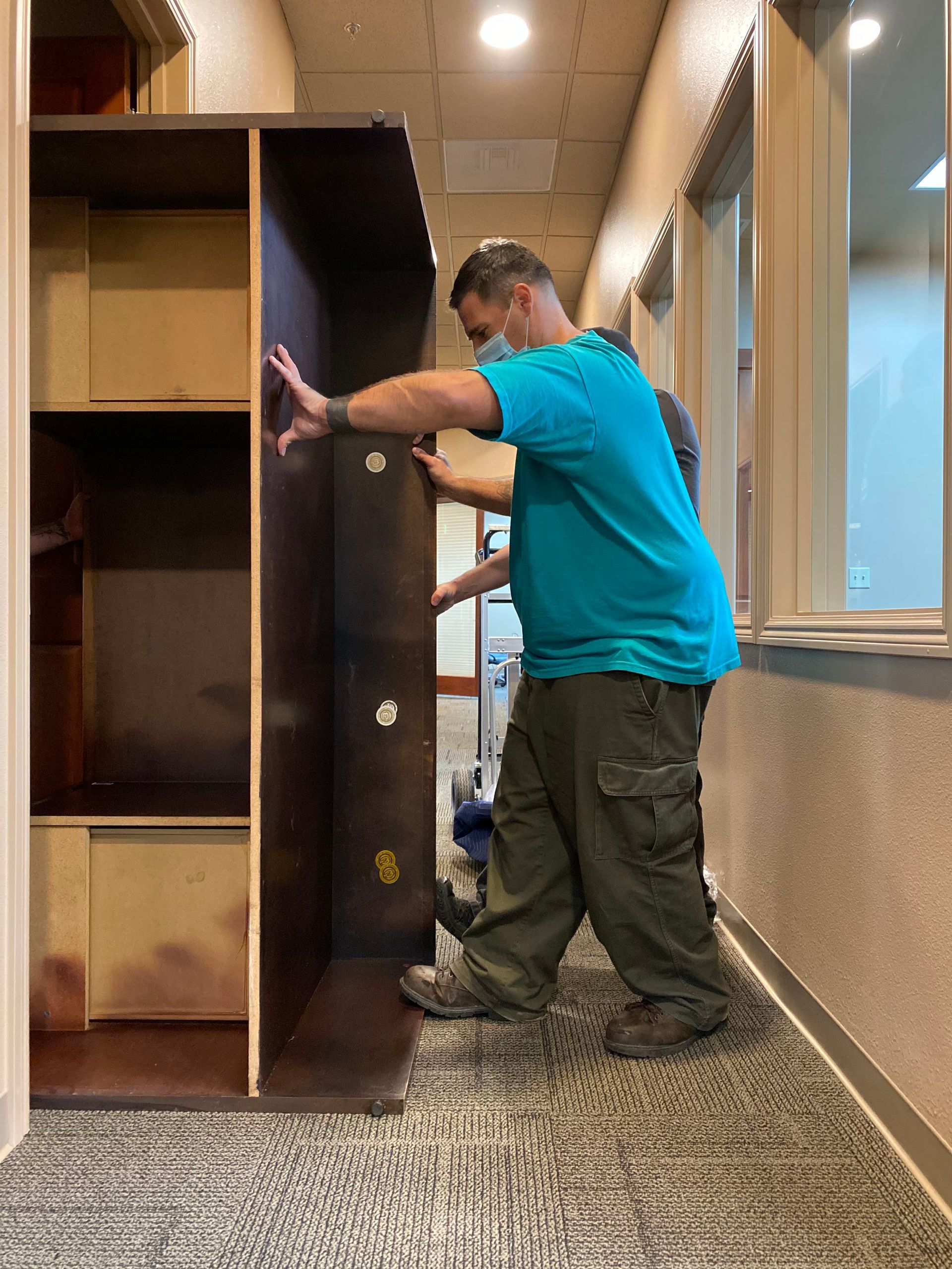 Person moving a tall dark cabinet down a hallway beside large windows