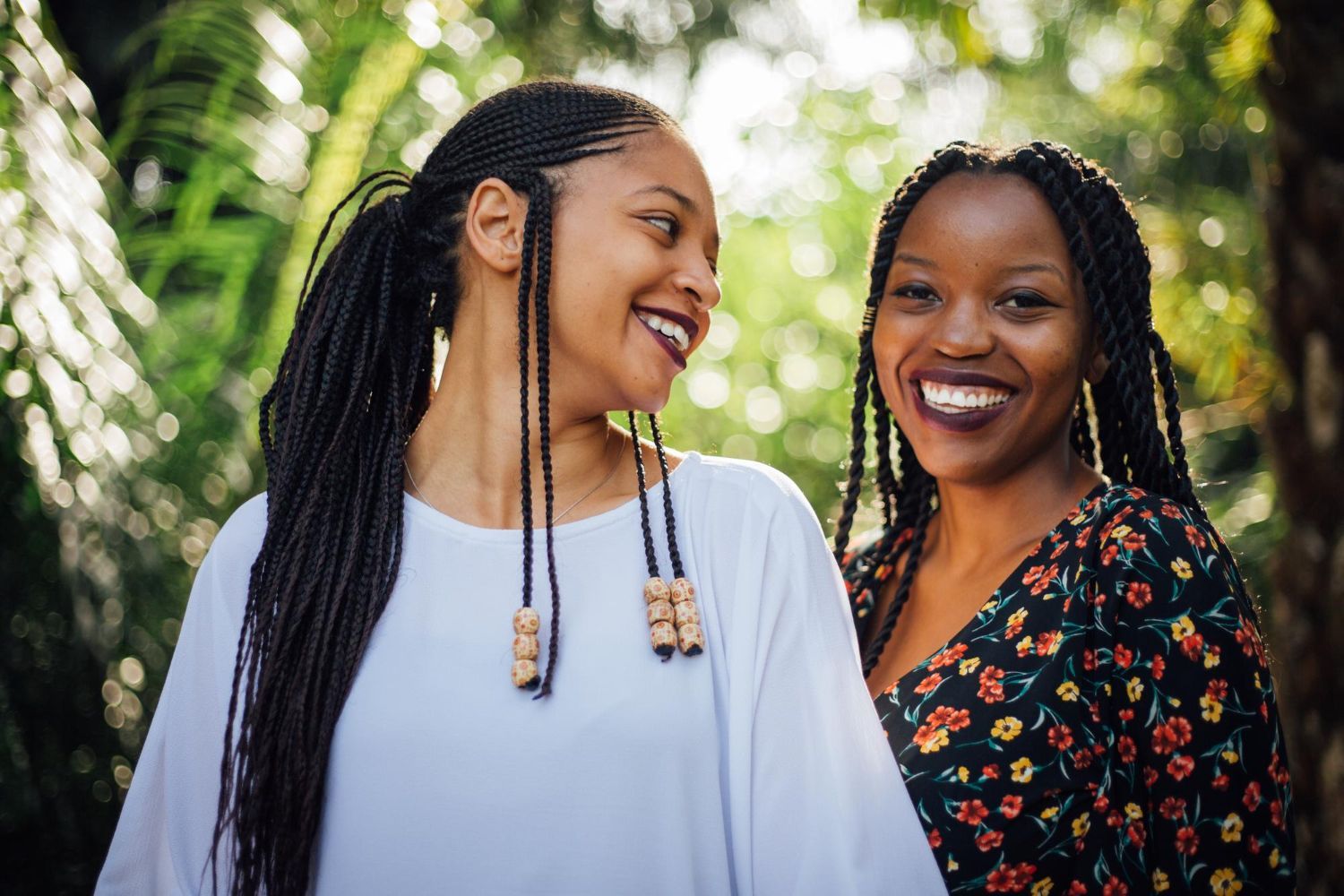 Duas mulheres negras sorrindo, ao ar livre. Uma usa uma camisa branca, a outra, um vestido florido.